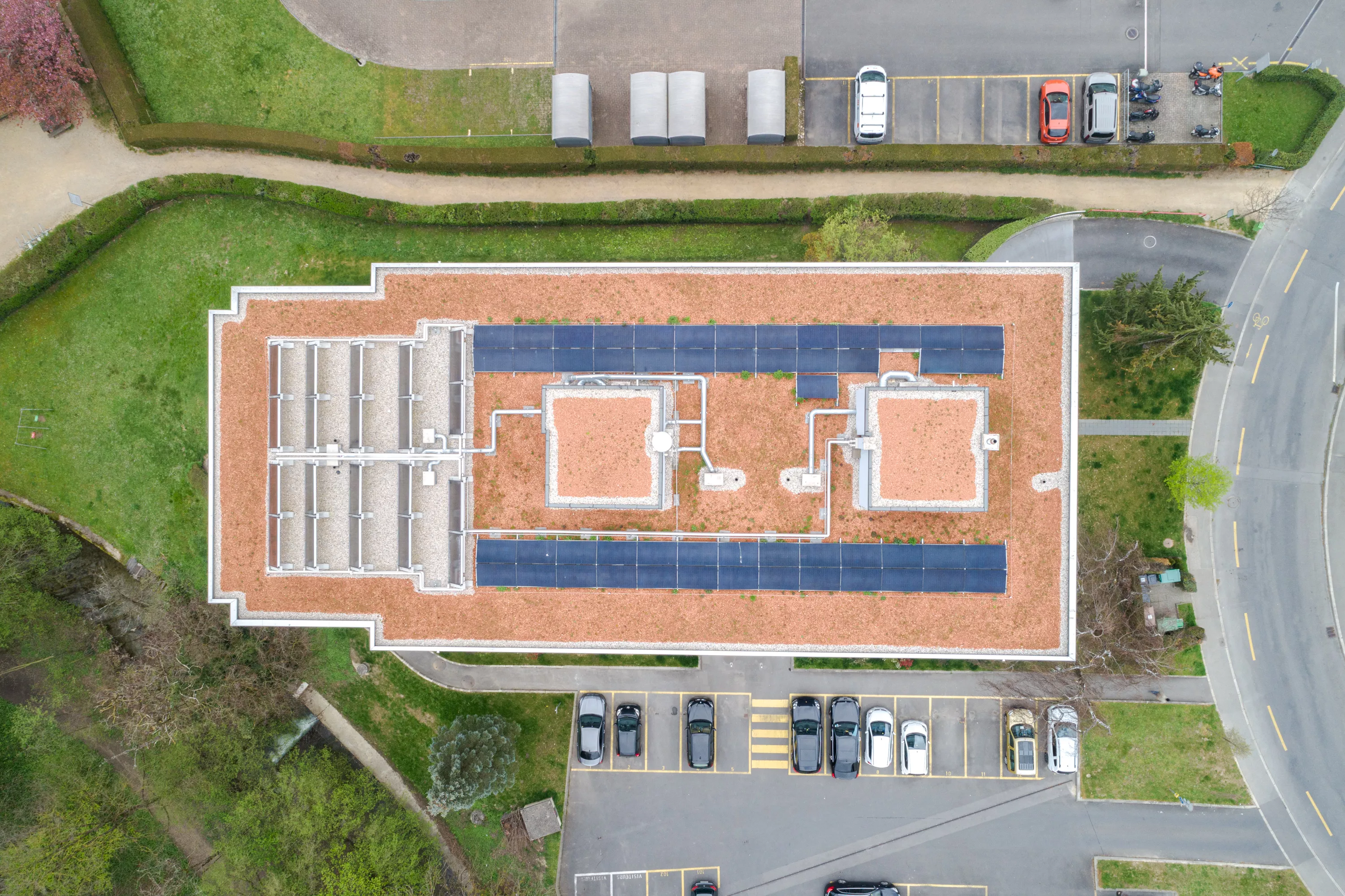 Image aérienne d'un complexe résidentiel récemment rénové, entouré de véhicules stationnés et de voies d'accès. La toiture est équipée de panneaux solaires, indiquant une mise à jour vers des solutions énergétiques durables.